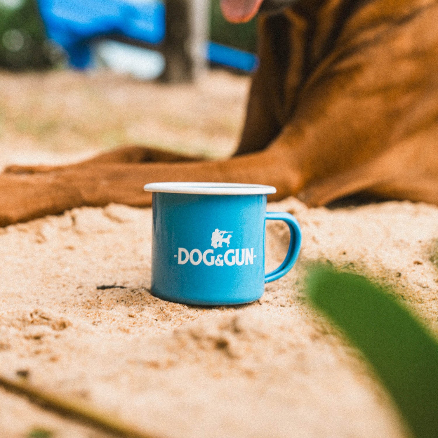 Blue enamel mug with 'DOG&GUN' branding next to a dog's paw on sandy ground
