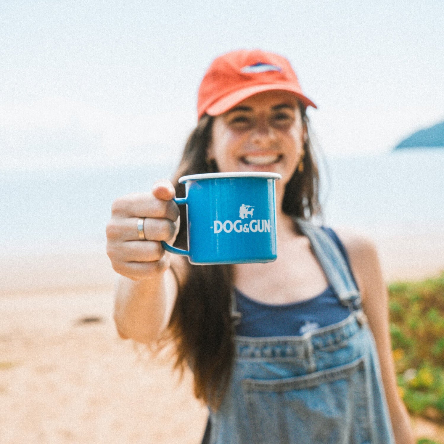Person holding a blue mug with 'Dog & Gun' branding on a beach.