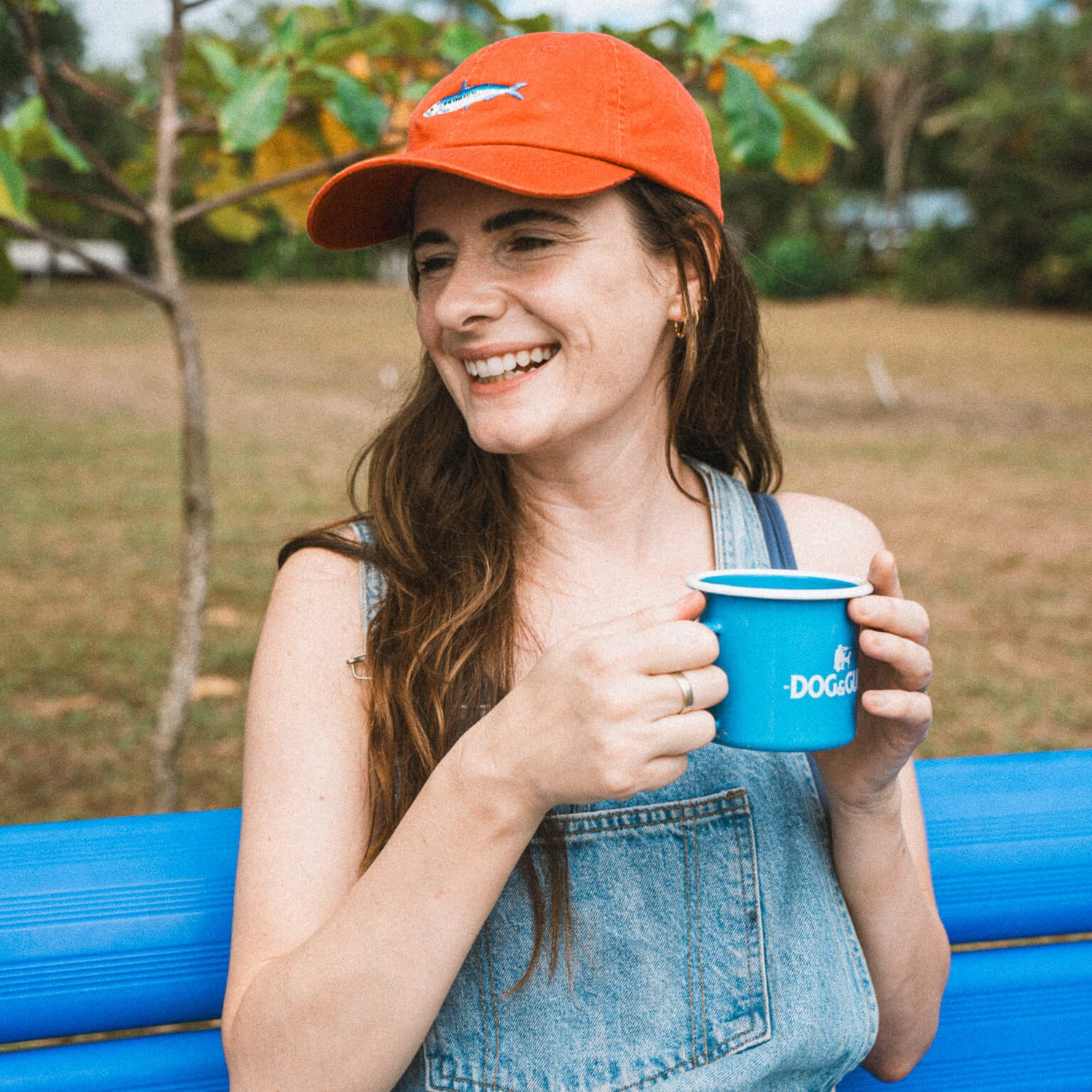 Woman holding a blue enamel mug with Dog & Gun on it. Outdoors with trees and grass in the background